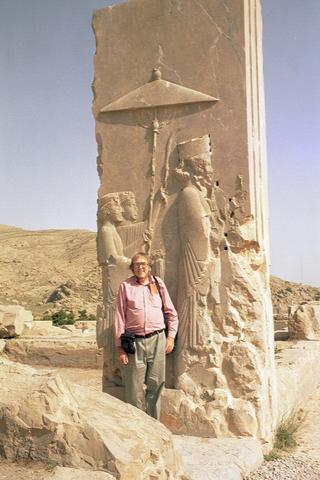 Full body portrait of Peter stood in front of an ancient monument, wearing salmon pink shirt and grey chinos