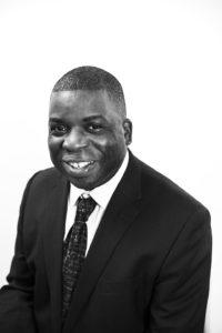 Black and white posed photo of man wearing dark jacket, white shirt and dark tie on white background