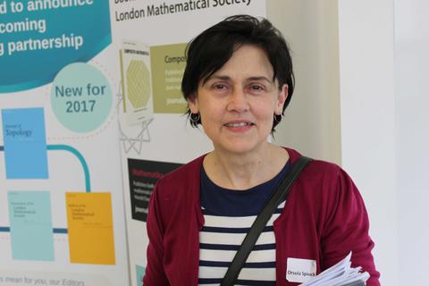 Photo of Orsola in front of maths poster, wearing red cardigan and black and white striped top