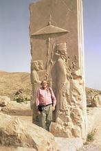 Full body portrait of Peter stood in front of an ancient monument, wearing salmon pink shirt and grey chinos